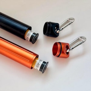 Op de afbeelding: Zwarte en oranje cilindrische containers met zilveren karabijnhaakclips. De containers zijn open en onthullen een geel en wit cilindrisch object binnenin. De tekst "RA 400" is zichtbaar op de witte cilinder.