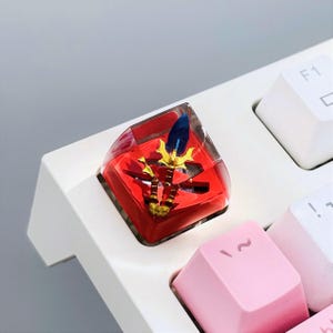 Puede incluir: Una tecla de teclado personalizada con una base roja y un diseño de espada dorada y azul, encapsulada en resina transparente. La tecla está en un teclado blanco, con teclas rosas y blancas visibles.