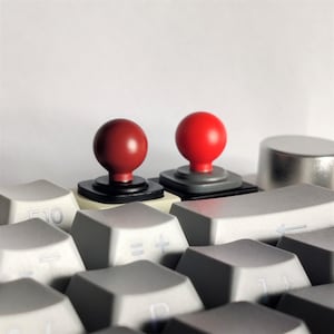 Puede incluir: Dos botones rojos de joystick en un teclado de computadora gris. Los botones están sobre bases negras y grises.