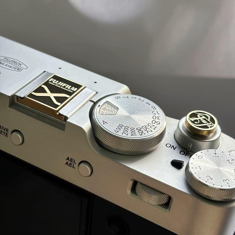May include: A silver camera with a black and gold Fujifilm X logo on the top. The camera has a silver dial with numbers from 1 to 8 and a silver button with the word "ON OFF" on it.