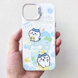 以下が含まれることがあります： 爽やかな夏の飲み物の漫画イラストが描かれた白い携帯電話ケース。ケースは「Lucky Summer」と「So Cool」の文字が入った青と白のデザインです。ケースには漫画のキャラクターとレモンのイラストも描かれています。