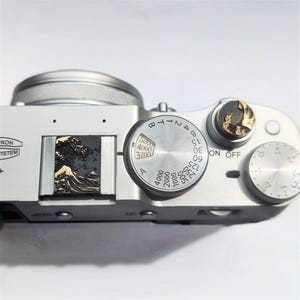 May include: A silver digital camera top plate featuring a circular shutter speed dial with numbers ranging from 3200 to 5000, a round exposure compensation dial, and a hot shoe cover embossed with a gold and black relief of a large ocean wave.