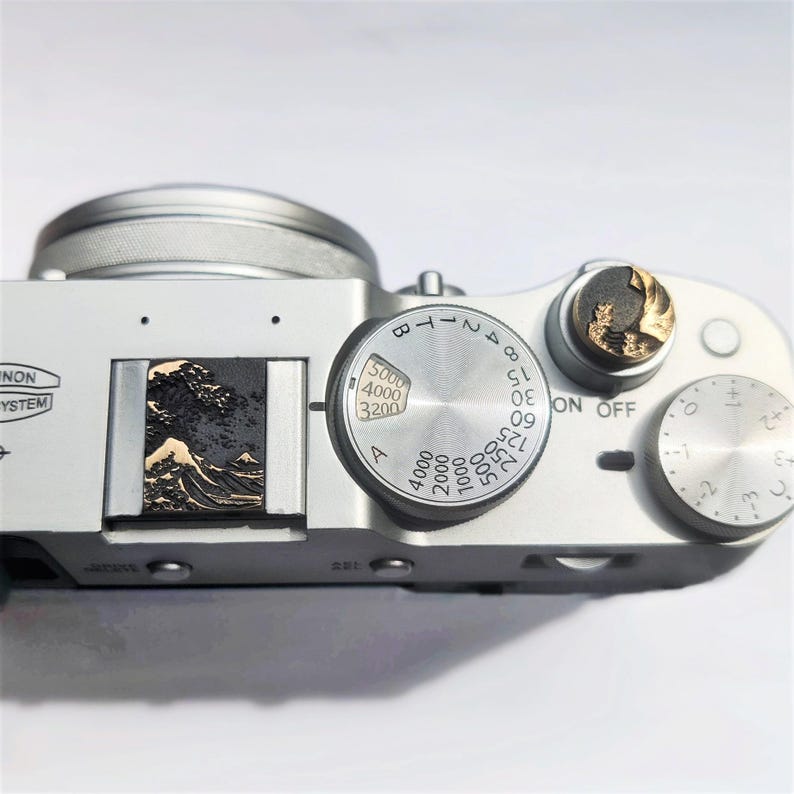 May include: A silver digital camera top plate featuring a circular shutter speed dial with numbers ranging from 3200 to 5000, a round exposure compensation dial, and a hot shoe cover embossed with a gold and black relief of a large ocean wave.