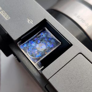 Könnte beinhalten: Nahaufnahme eines Kameraobjektivs mit einer blauen und schwarzen Glitzerüberlagerung. Das Objektiv ist mit "F3.6/16-50" beschriftet.