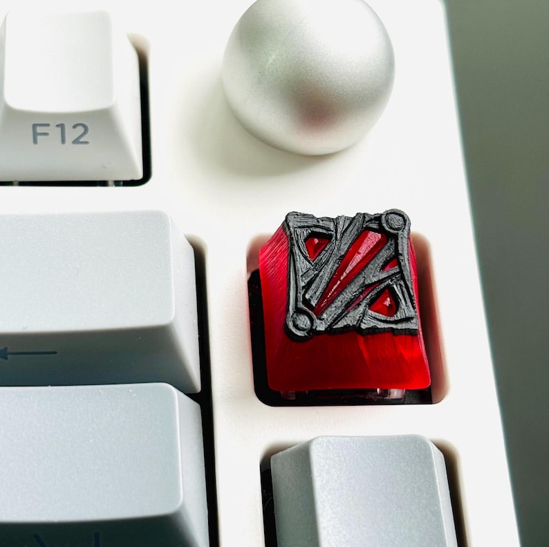 May include: A red mechanical keyboard keycap with a black and gray design featuring a stylized logo. The keycap is on a white keyboard.