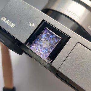 May include: A silver camera with a black lens and a clear plastic cover over the viewfinder. The viewfinder is filled with blue and silver glitter. The camera has the text "a 6000" on the front.