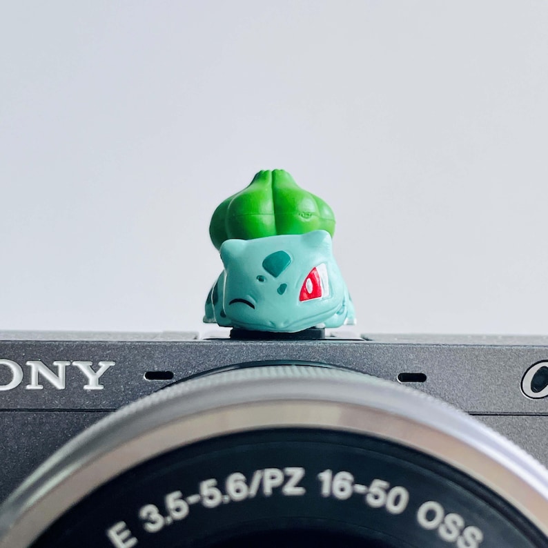 May include: A small, light blue Bulbasaur figurine with a green bulb on its back, resting on a gray camera. The figurine has red accents on its face and is set against a white background. The camera lens has the text "E 3.5-5.6/PZ 16-50 OSS".