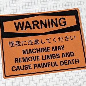 Puede incluir: Señal de advertencia naranja y negra con el texto "WARNING" en letras negras grandes. La señal también incluye el texto "怪我に注意してください MACHINE MAY REMOVE LIMBS AND CAUSE PAINFUL DEATH" en letras negras.