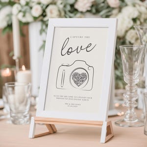 May include: White framed sign with a black and white illustration of a camera with a heart containing a QR code. The sign reads "Capture the love" and "Scan the QR code to upload your photos to our online album". The sign also includes the names "Mrs & Mrs Jones" and the date "01.04.2021".