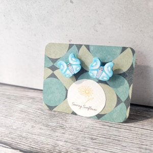 Puede incluir: Un par de pendientes de mariposa en color azul claro con detalles blancos y centro oscuro, sujetos a una tarjeta de presentación rectangular con un patrón geométrico en tonos verdes y grises y un logotipo circular que dice Soaring Sunflower.