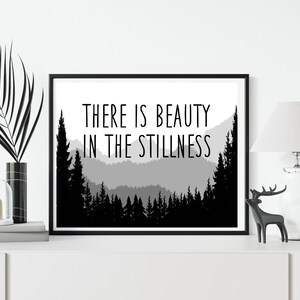 May include: Black and white print with a mountain range silhouette and the text "THERE IS BEAUTY IN THE STILLNESS".