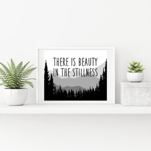 May include: Black and white print with a silhouette of a mountain range and trees. The text "THERE IS BEAUTY IN THE STILLNESS" is printed in black.
