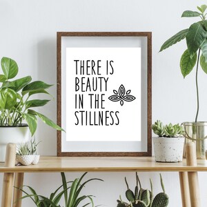 Puede incluir: Una impresi&oacute;n enmarcada con fondo blanco y texto negro que dice "There is beauty in the stillness" con un gr&aacute;fico de flor de loto estilizado. La impresi&oacute;n est&aacute; sobre una mesa de madera con plantas en macetas.