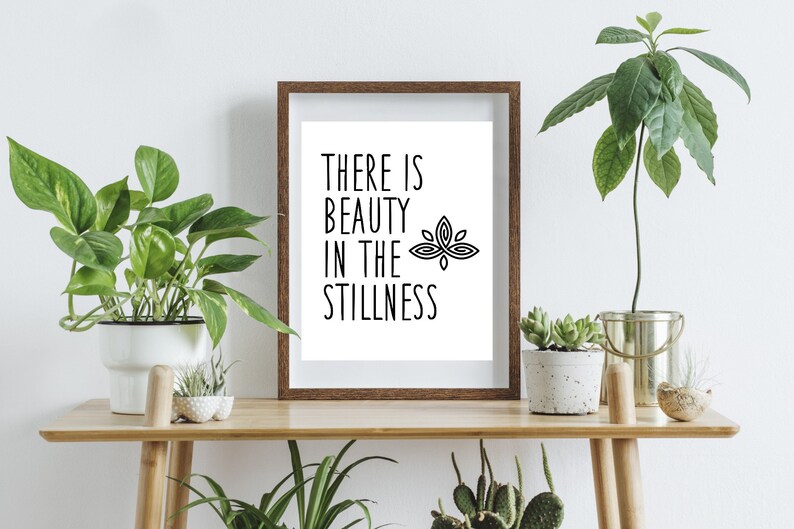 Puede incluir: Una impresi&oacute;n enmarcada con fondo blanco y texto negro que dice "There is beauty in the stillness" con un gr&aacute;fico de flor de loto estilizado. La impresi&oacute;n est&aacute; sobre una mesa de madera con plantas en macetas.