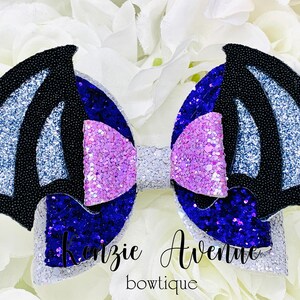 May include: A purple, blue, and silver glitter bow with black bat wings. The bow is decorated with a white bow in the center. The text "Kenzie Avenue boutique" is written below the bow.