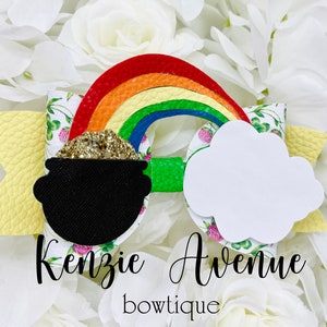 Op de afbeelding: Een gele, groene en witte strik met een regenboog, een pot met goud en een witte wolk. De strik is versierd met klavertjesvier en de tekst "Kenzie Avenue boutique".