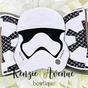 以下が含まれることがあります： ストームトルーパーのヘルメットのデザインが施された白、黒、赤のボウ。ボウは黒のグリッターと「Kenzie Avenue bowtique」の文字で飾られています。