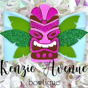 Peut inclure: Une tête de tiki rose et marron avec des feuilles vertes à paillettes sur un fond bleu clair. Le texte "Kenzie Avenue bowtique" est sous la tête de tiki.