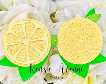 Può includere: Un fiocco per capelli giallo glitterato a forma di limone con foglie verdi. Il fiocco ha un'incisione a forma di fetta di limone al centro. Il testo "Kenzie Avenue bowtique" è scritto sotto il fiocco.