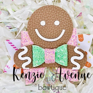 May include: A gingerbread man-shaped hair bow with a pink and green glitter bow, white icing details, and the text "Kenzie Avenue bowtique".