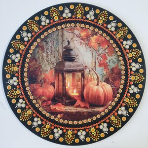 May include: A circular wall plaque featuring a central image of a rustic lantern with lit candles, surrounded by orange pumpkins and autumn leaves. The border is decorated with a symmetrical pattern of hand-painted dots in shades of orange, yellow, and white.