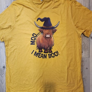 Puede incluir: Una camiseta amarilla con una imagen de dibujos animados de una vaca de las Tierras Altas con un sombrero de bruja. El texto "Moo... I mean Boo!" está impreso debajo de la vaca.