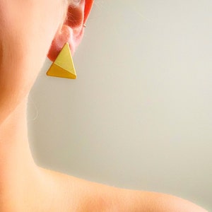 Könnte beinhalten: Ein Paar goldene Dreiecksohrringe. Die Ohrringe bestehen aus einem massiven Stück Metall und haben eine geometrische Form.