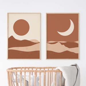 May include: Two framed abstract art prints. One features a large brown sun over brown and beige layered hills. The other print shows a white crescent moon over similar hills. Both are in light wood frames. A wicker baby bassinet is in the foreground.