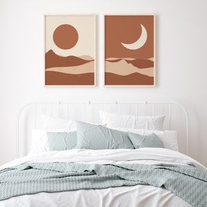 May include: Two framed art prints depicting a desert landscape with a sun and moon. The artwork features a warm color palette of brown, beige, and cream. The prints are displayed above a bed with white linens, blue pillows, and a gray blanket.