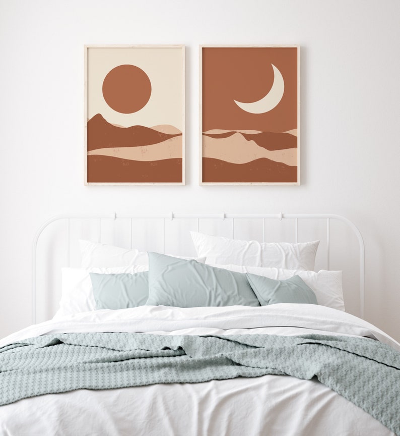 May include: Two framed art prints depicting a desert landscape with a sun and moon. The artwork features a warm color palette of brown, beige, and cream. The prints are displayed above a bed with white linens, blue pillows, and a gray blanket.