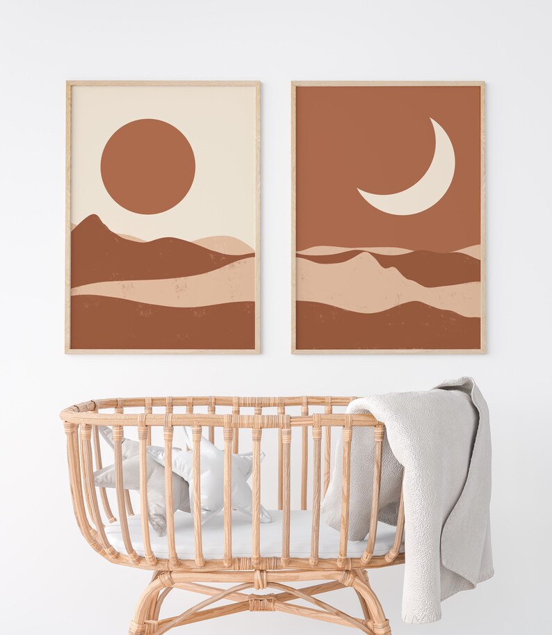 May include: Two framed abstract art prints. One features a large brown sun over brown and beige layered hills. The other print shows a white crescent moon over similar hills. Both are in light wood frames. A wicker baby bassinet is in the foreground.