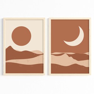 May include: Two framed art prints with a minimalist desert landscape theme. One features a rust-colored sun over brown and beige hills against a cream background. The other shows a cream crescent moon over similar hills against a rust background.