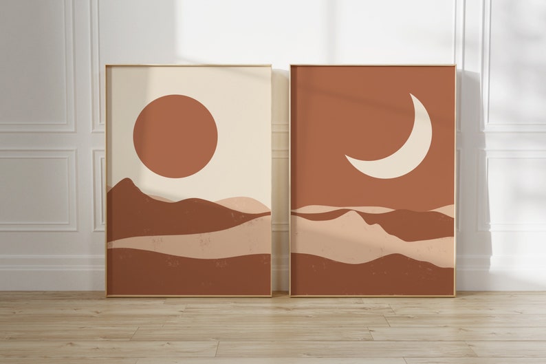 May include: Two framed art prints with a minimalist desert landscape design. One features a large orange sun over brown hills against a cream background. The other shows a white crescent moon over similar hills.