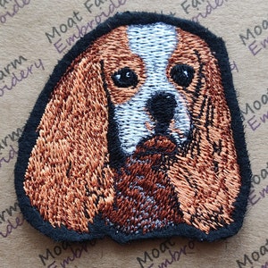 May include: Embroidered patch of a Cavalier King Charles Spaniel dog with brown and white fur. The dog is looking directly at the viewer.