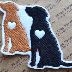 May include: Two embroidered dog silhouettes, one brown and one black, are sitting side-by-side. Each dog has a white heart on its chest.