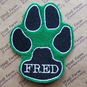 May include: Green paw print with black paw pads and a white outline. The name FRED is embroidered in white on the bottom of the paw print.