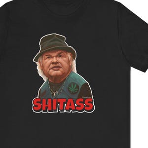 May include: Black t-shirt with a graphic of a man wearing a hat and vest, and the word "SHITASS" in red and white. The man's portrait includes a cannabis leaf on his vest.