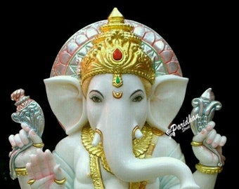 May include: White marble statue of Ganesha, the Hindu god of wisdom and prosperity, sitting with his trunk curled and holding a sweet in his hand. He is adorned with gold jewelry and a crown. The statue is on a white marble base with gold trim.
