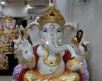 May include: A white marble statue of the deity Ganesha seated on a gold-painted throne. The figure features gold-leaf detailing, a red garment, and a crown with a red stone. Two small brown mouse figures are positioned at the base of the statue.