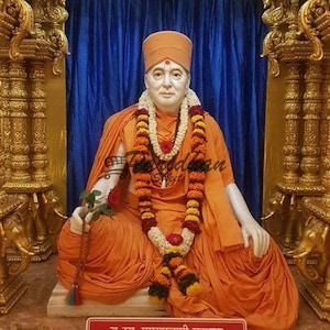 May include: A statue of a man wearing an orange robe and a garland of flowers. He is sitting on a throne with a blue curtain behind him. The statue is in a golden temple with intricate carvings. The text on the plaque reads "Br. Sv. Pramukhswami Maharaj".