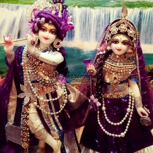 May include: A pair of intricately decorated figurines of Krishna and Radha, dressed in purple and gold garments with pearl necklaces and elaborate headwear. The figurines are standing in front of a waterfall backdrop.