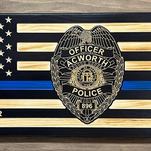 May include: Wooden American flag with a blue line, a police badge, and the name "JAMES WALKER" with the years "2018-2024". The badge reads "OFFICER ACWORTH POLICE". The flag has a black and natural wood finish.