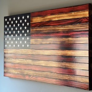 May include: Wooden American flag wall art. The flag features a dark blue canton with white stars and red and natural wood stripes. The wood has a distressed finish, giving it a rustic appearance.