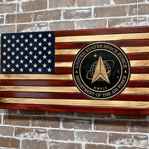 May include: Wooden American flag with the United States Space Force emblem. The flag features the traditional red and white stripes, a blue field with stars, and a circular emblem with text and a logo. The flag is mounted on a brick wall.