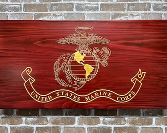 May include: A wooden sign with a red background and a gold United States Marine Corps emblem. The emblem features a globe, an anchor, and a ribbon with the words "United States Marine Corps".