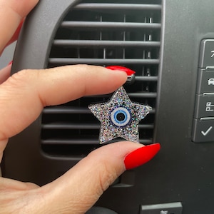 May include: A silver star-shaped car air freshener with a blue evil eye charm. The air freshener is hanging from a car vent.