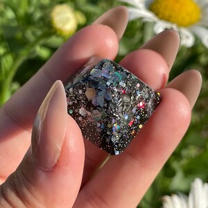 May include: A black cube with iridescent glitter embedded in the resin. The glitter is a mix of silver, gold, and holographic colors.