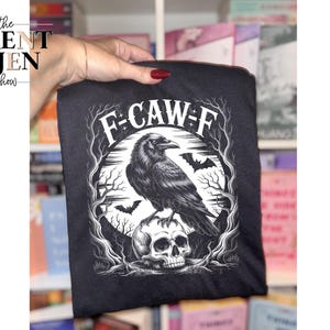 May include: Black t-shirt featuring a detailed illustration of a raven perched on a skull, surrounded by a circular design with bats and tree branches. The text "F-CAW-F" is arched above the raven. The Brent & Jen Show logo is in the upper left corner.