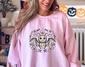 May include: A light pink sweatshirt featuring a cute bat design with a crescent moon and floral accents. The bat has large eyes and outstretched wings. The text "the BRENT & JEN show" is at the bottom left.
