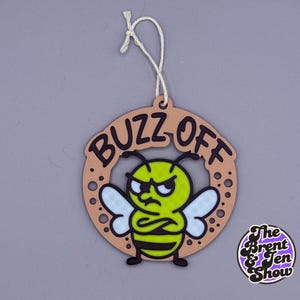 May include: A round, tan-colored ornament with the words "BUZZ OFF" in brown lettering. The ornament features a cartoon bee with an angry expression. The bee is yellow, black, and white. A white string is attached for hanging.